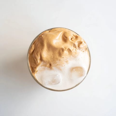 Two glasses of Dalgona coffee feature cloud-like whipped coffee floating over iced milk in a cafe-style presentation.  