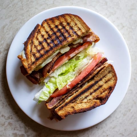A golden, buttery Vegan Bacon BLT Grilled Cheese rests on a cutting board, cut in half to reveal layers of fresh lettuce and tomato.