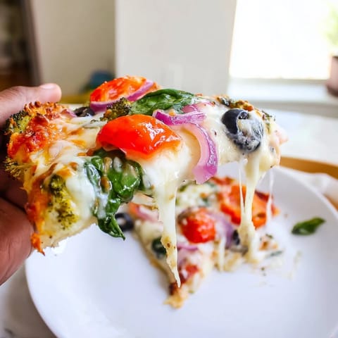 Slice of low-carb Broccoli Chicken Crust Pizza on a wooden board, revealing colorful toppings of red bell peppers, black olives, and savory Italian herbs.